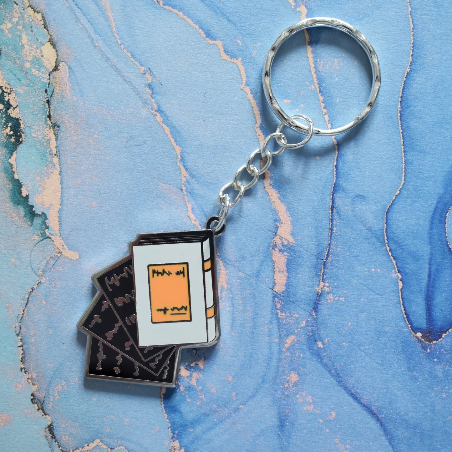 A stainless steel ring keyrings at an angle across the centre of the image. With a silver metal enamel piece on the end. Design is book facing left in grey with black pages falling out. Background of photo is swirly blue.