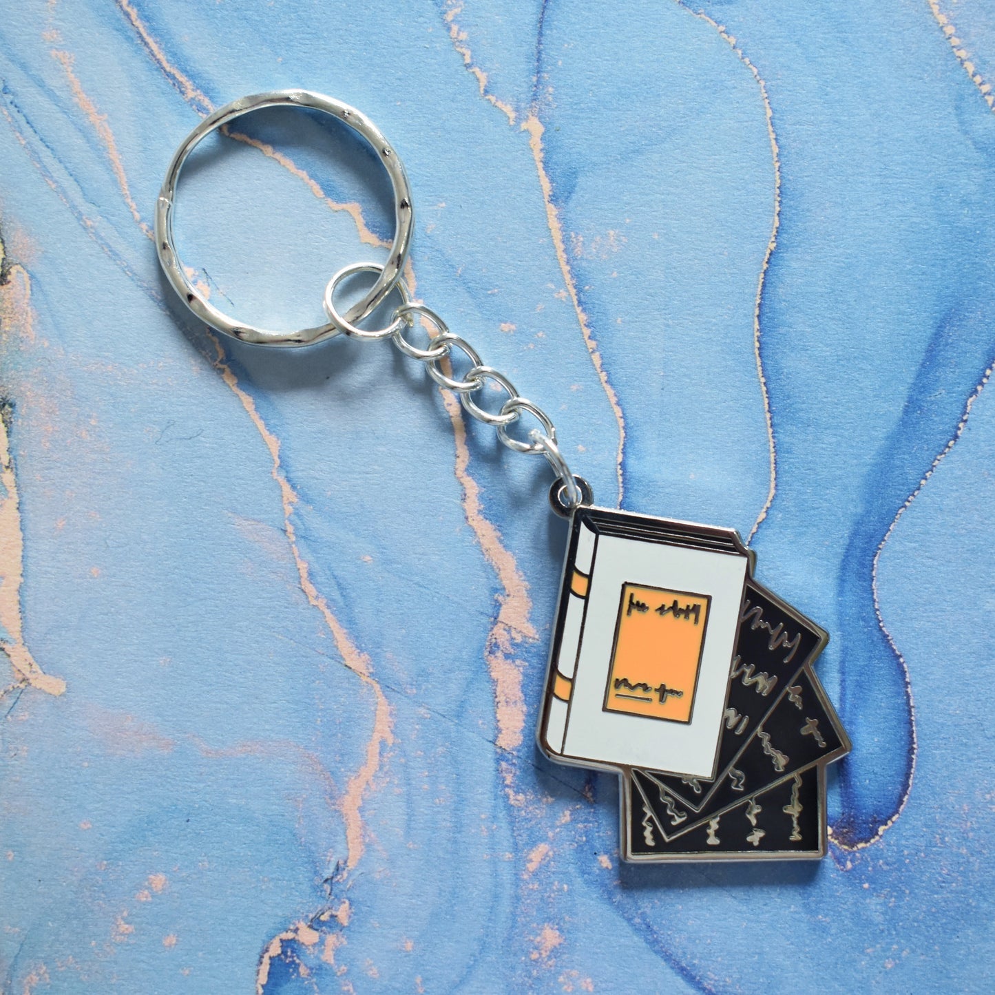 A stainless steel ring keyrings at an angle across the centre of the image. With a silver metal enamel piece on the end. Design is book facing right in grey with black pages falling out. Background of photo is swirly blue.