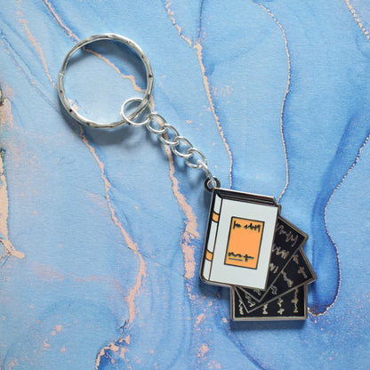 A stainless steel ring keyrings at an angle across the centre of the image. With a silver metal enamel piece on the end. Design is book facing right in grey with black pages falling out. Background of photo is swirly blue.