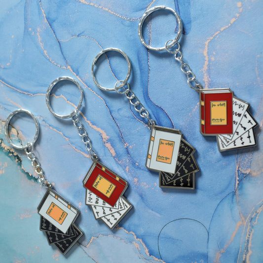 4 stainless steel ring keyrings in a line at an angle across the centre of the image. Each with a silver metal enamel piece on the end. Left to right, book facing left in grey with black pages falling out, book facing left in red with white pages falling out, book facing right in grey with black pages falling out and book facing right in red with white pages falling out. Background of photo is swirly blue.