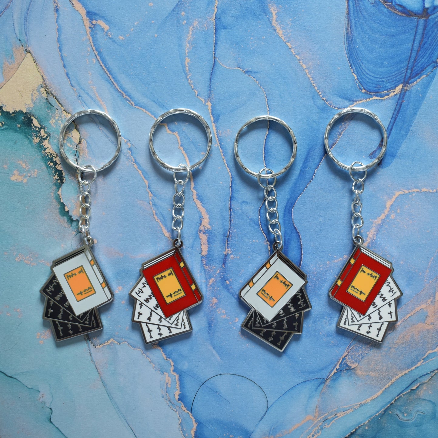 4 stainless steel ring keyrings in a line across the centre of the image. Each with a silver metal enamel piece on the end. Left to right, book facing left in grey with black pages falling out, book facing left in red with white pages falling out, book facing right in grey with black pages falling out and book facing right in red with white pages falling out. Background of photo is swirly blue.
