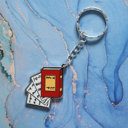 A stainless steel ring keyrings at an angle across the centre of the image. With a silver metal enamel piece on the end. Design is book facing left in red with white pages falling out. Background of photo is swirly blue.