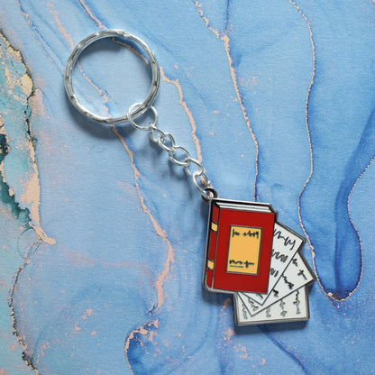 A stainless steel ring keyrings at an angle across the centre of the image. With a silver metal enamel piece on the end. Design is book facing right in red with white pages falling out. Background of photo is swirly blue.