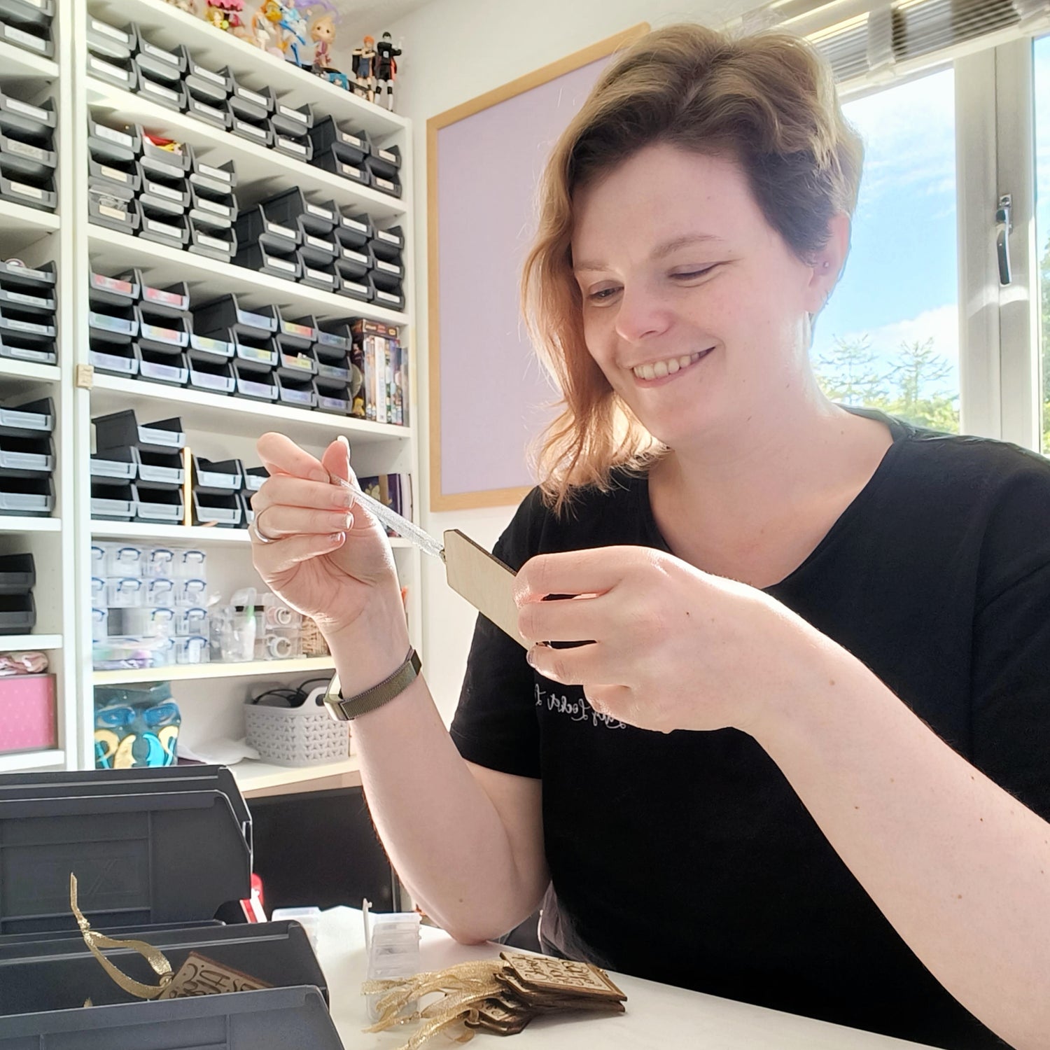 Photograph of a white person with brown hair that is short on one side & long on the other. They are holding a wooden ornagment in their hands and looking at it. More wooden ornaments are on the table in from of them Shelves filled with labelled grey tubs, a purple pin board and a window are in the background.