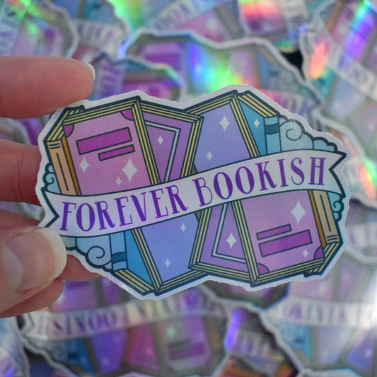 White hand holding sticker. Sticker design is pastel coloured books and pastel blue curly clouds running across the top and bottom of a white banner that reads forever bookish. Sticker has a white border all around the design. Background is blurry/distance of the same stickers scattered.