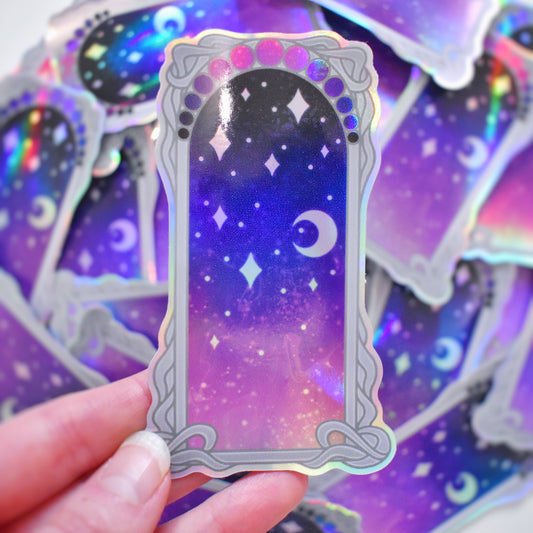White hand holding sticker. Sticker design is a grey curly frame with a gradient of circles across the top, from dark navy to blue to pinkish purple. Centre of the design is a dark navy to blue to pinkish purple gradient with white/hazy stars and moon. Background is blurry/distance of same stickers.