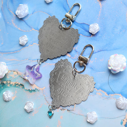 2 keyrings with stainless steel engraved heart and book charm that reads happily ever after at diagonal in the centre of image. Left keyring has a lilac glittery cabachon star attached to charm. Right keyring has a small silver and gem pale blue heart attached to charm. On a pale blue/teal watercolour background with white rose cabachons scattered around.