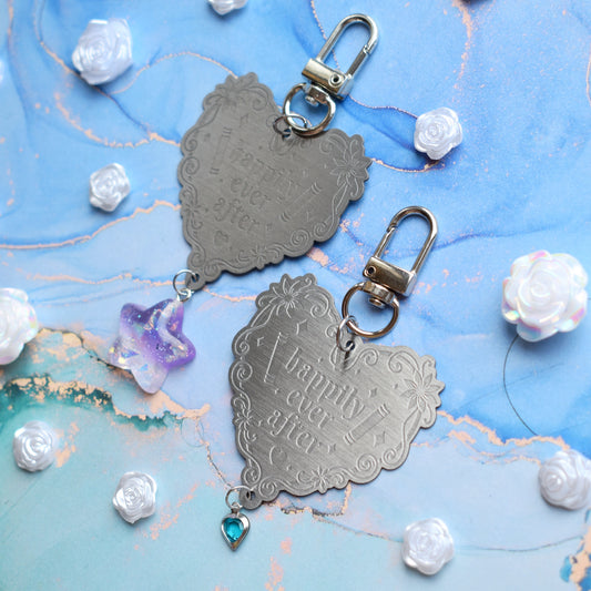 2 keyrings with stainless steel engraved heart and book charm that reads happily ever after at diagonal in the centre of image. Left keyring has a lilac glittery cabachon star attached to charm. Right keyring has a small silver and gem pale blue heart attached to charm. On a pale blue/teal watercolour background with white rose cabachons scattered around.