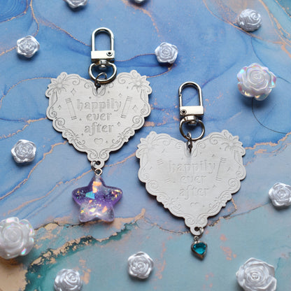 2 keyrings with stainless steel engraved heart and book charm that reads happily ever after in the centre of image. Left keyring has a lilac glittery cabachon star attached to charm. Right keyring has a small silver and gem pale blue heart attached to charm. On a pale blue/teal watercolour background with white rose cabachons scattered around.