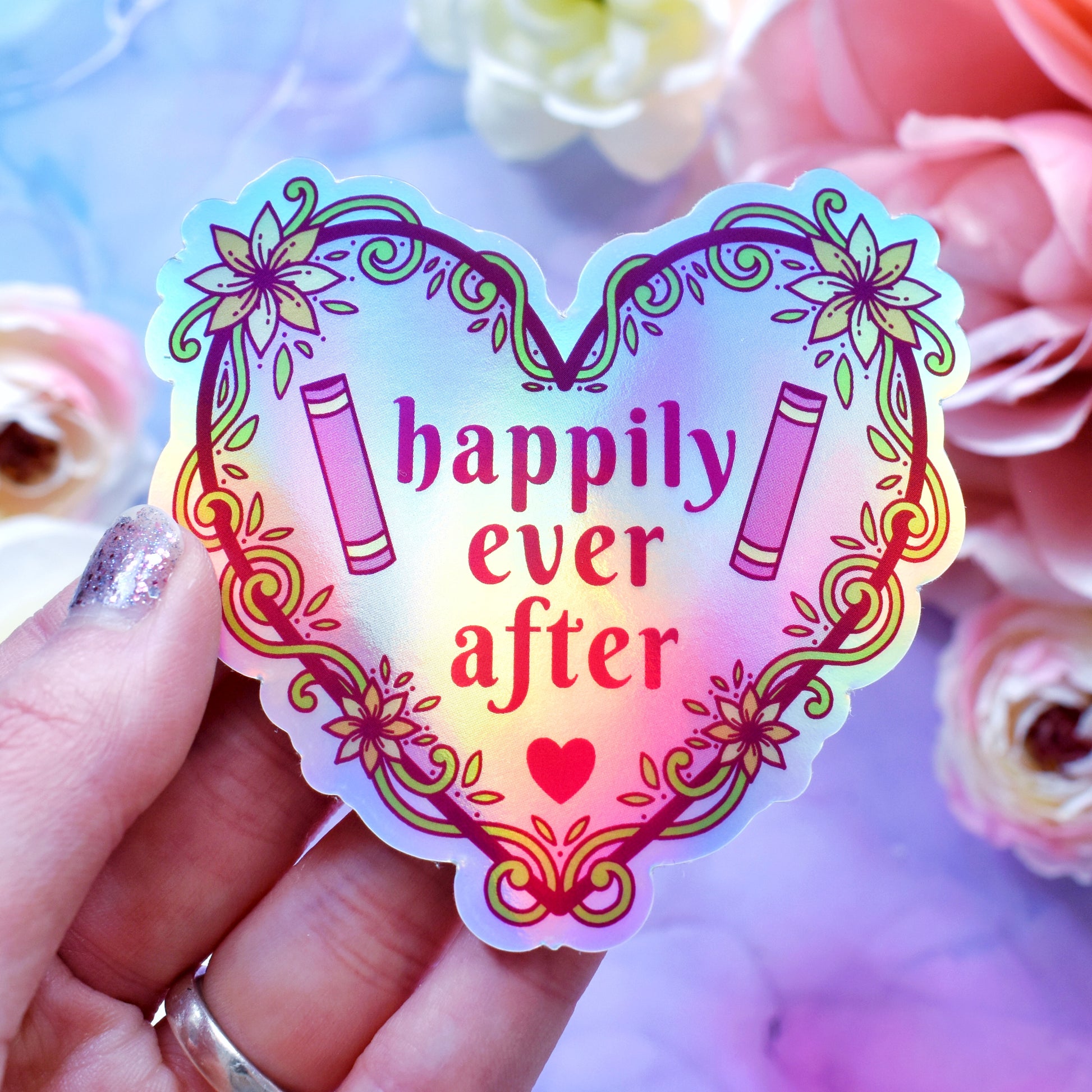 Pink heart-shaped bookish holographic sticker design features text 'happily ever after' with a floral and swirly outline with book spines. Held by a white hand above a watercolour style background in blue, pink and purples with flowers around the edge of the photo.