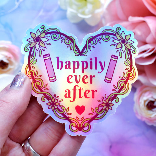 Pink heart-shaped bookish holographic sticker design features text 'happily ever after' with a floral and swirly outline with book spines. Held by a white hand above a watercolour style background in blue, pink and purples with flowers around the edge of the photo.