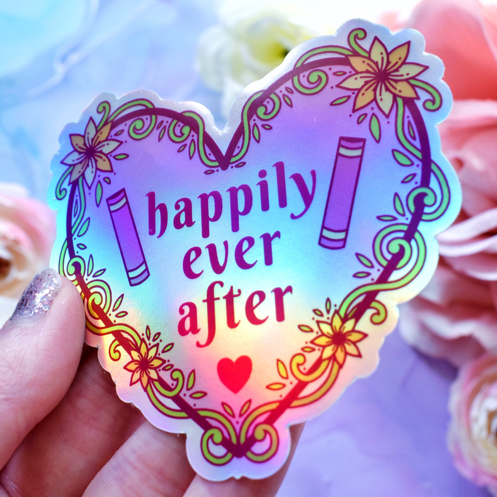 Pink heart-shaped bookish holographic sticker design features text 'happily ever after' with a floral and swirly outline with book spines. Held at an angle by a white hand above a watercolour style background in blue, pink and purples with flowers around the edge of the photo.