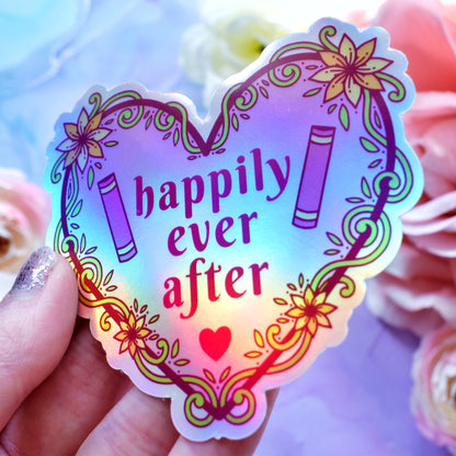 Pink heart-shaped bookish holographic sticker design features text 'happily ever after' with a floral and swirly outline with book spines. Held at an angle by a white hand above a watercolour style background in blue, pink and purples with flowers around the edge of the photo.