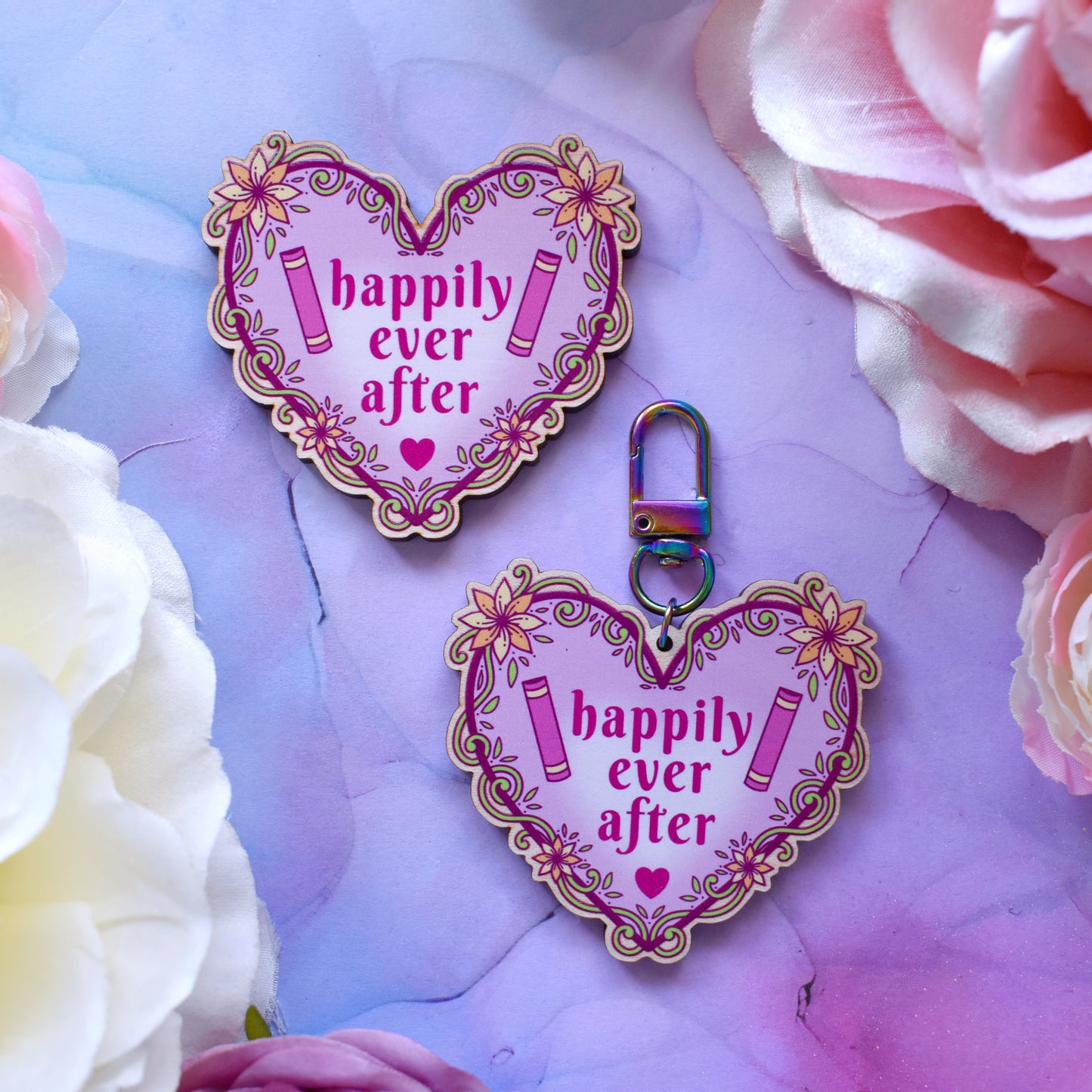 2 Pink heart-shaped bookish wooden products, charm and keyring version, design features text 'happily ever after' with a floral and swirly outline with book spines. Products are placed on a watercolour style background in blue, pink and purples with flowers around the edge of the photo.