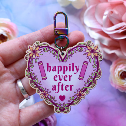 Pink heart-shaped bookish keyring version, design features text 'happily ever after' with a floral and swirly outline with book spines. Keyring held by a white hand above a watercolour style background in blue,pink and purples with flowers around the edge of the photo.