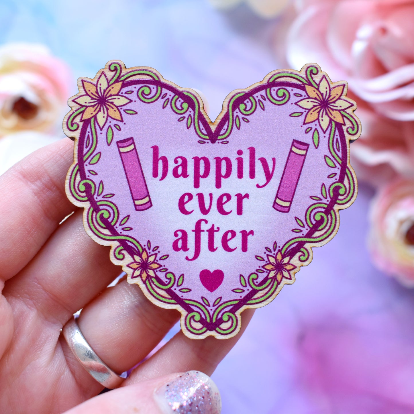 Pink heart-shaped bookish pin/magnet version, design features text 'happily ever after' with a floral and swirly outline with book spines. Pin/magnet held by a white hand above a watercolour style background in blue, pink and purples with flowers around the edge of the photo.