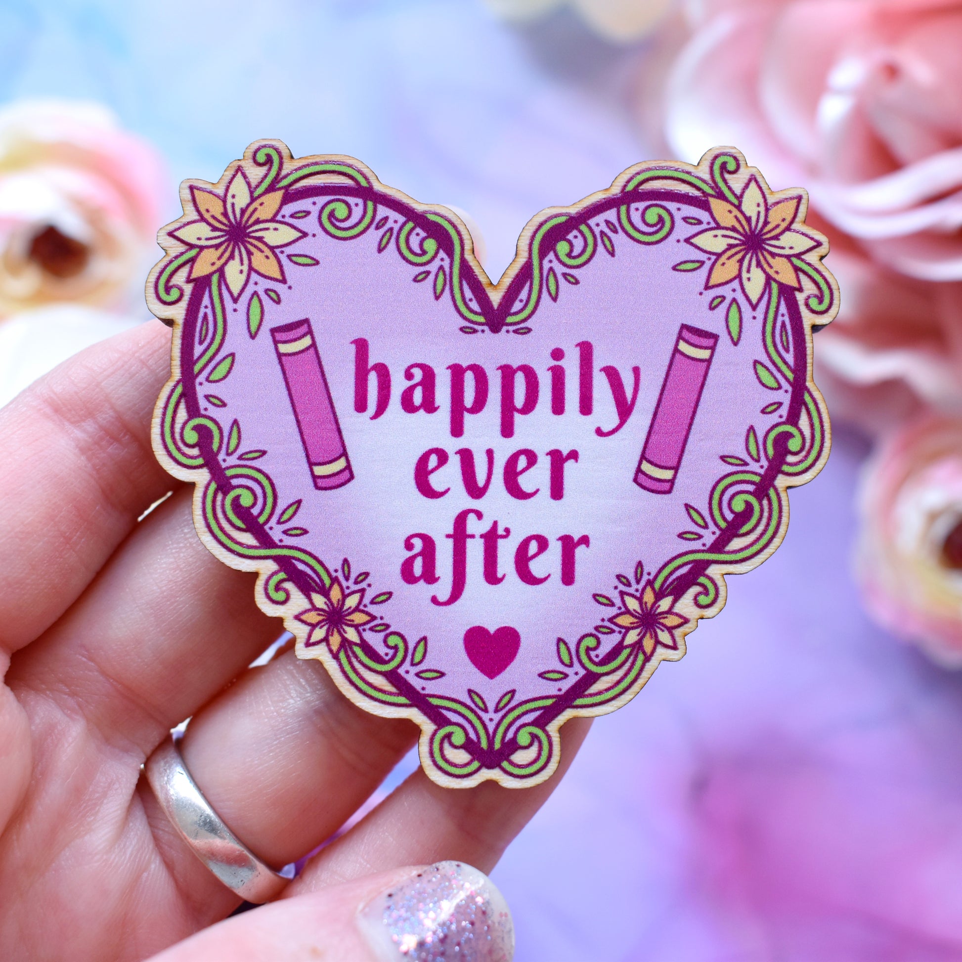 Pink heart-shaped bookish pin/magnet version, design features text 'happily ever after' with a floral and swirly outline with book spines. Pin/magnet held by a white hand above a watercolour style background in blue, pink and purples with flowers around the edge of the photo.
