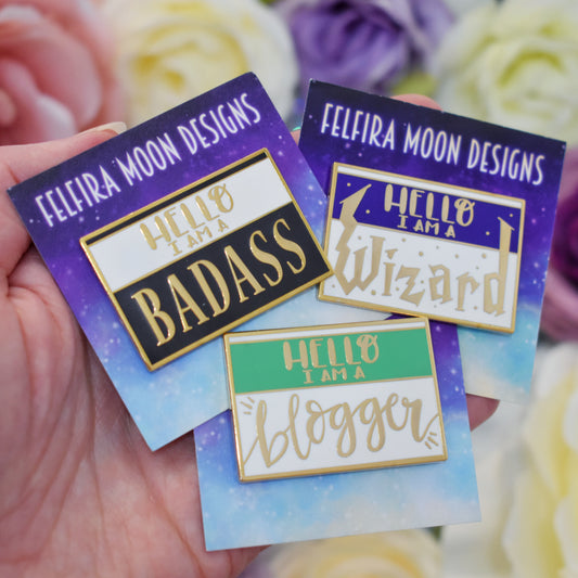 White handing holding 3 gold metal enamel pins on galaxy backing cards with felfira moon designs on. Far left pin is black and white with hello i am a badass written on it. Far right pin is purple and white with hello i am a wizard written on it. Bottom middle pin is green and white with hello i am a blogger writte on it. background is blurry white, pink and purple fake flowers.
