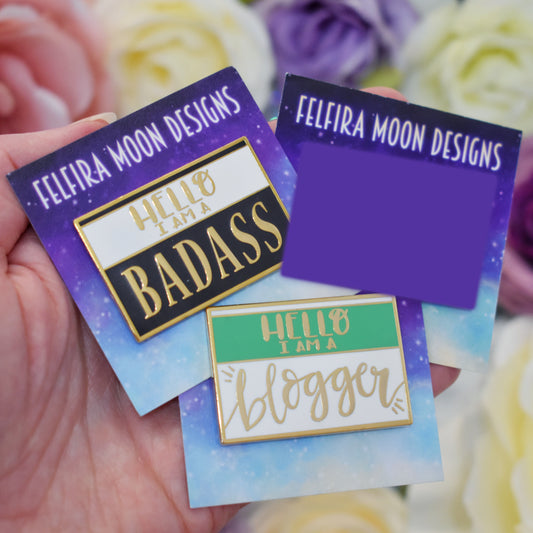 White handing holding 3 gold metal enamel pins on galaxy backing cards with felfira moon designs on. Far left pin is black and white with hello i am a badass written on it. Far right pin is blocked out by a purple rectangle as it is no longer sold. Bottom middle pin is green and white with hello i am a blogger writte on it. background is blurry white, pink and purple fake flowers.