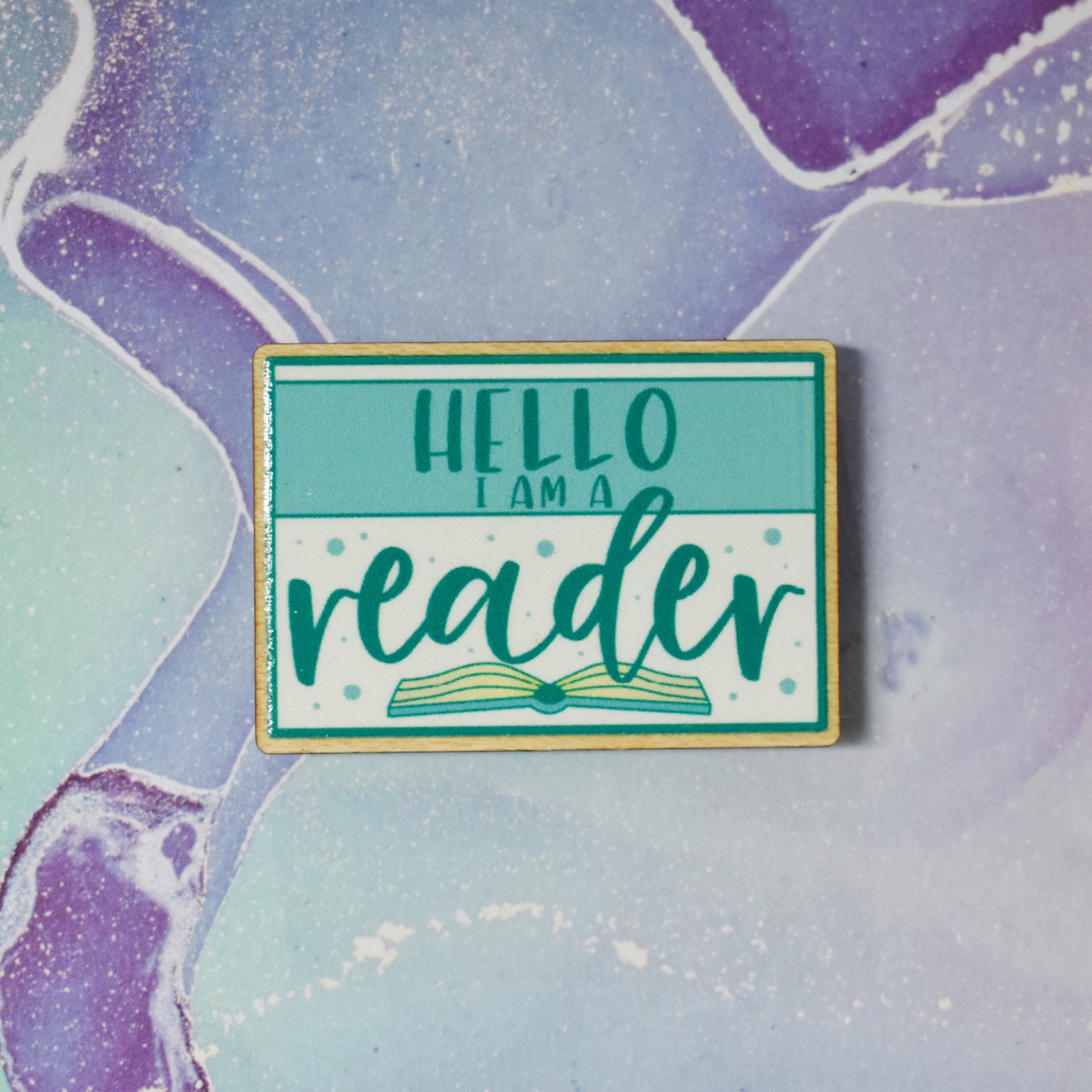 Wooden pin in centre of photo. Design features a teal thin border around the edge, with a lighter teal band across the design, with some white above and below the band. Hello I am a is written in a sans serif font across the tela band. Reader is written in the larger botton white space with an open book beneath. Background is soft watercolour teal and purple style.