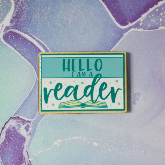 Wooden pin in centre of photo. Design features a teal thin border around the edge, with a lighter teal band across the design, with some white above and below the band. Hello I am a is written in a sans serif font across the tela band. Reader is written in the larger botton white space with an open book beneath. Background is soft watercolour teal and purple style.