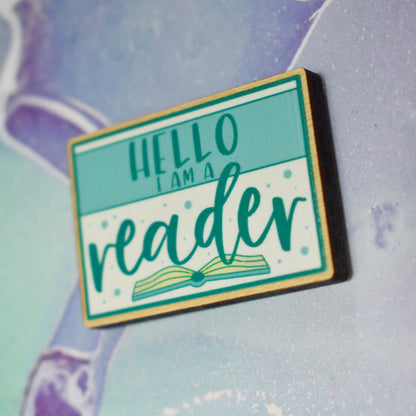 Close up of wooden pin in centre of photo at an angle, which shows the cut edge of the pin which is darker than the wood visable on the front. Design features a teal thin border around the edge, with a lighter teal band across the design, with some white above and below the band. Hello I am a is written in a sans serif font across the tela band. Reader is written in the larger botton white space with an open book beneath. Background is soft watercolour teal and purple style.