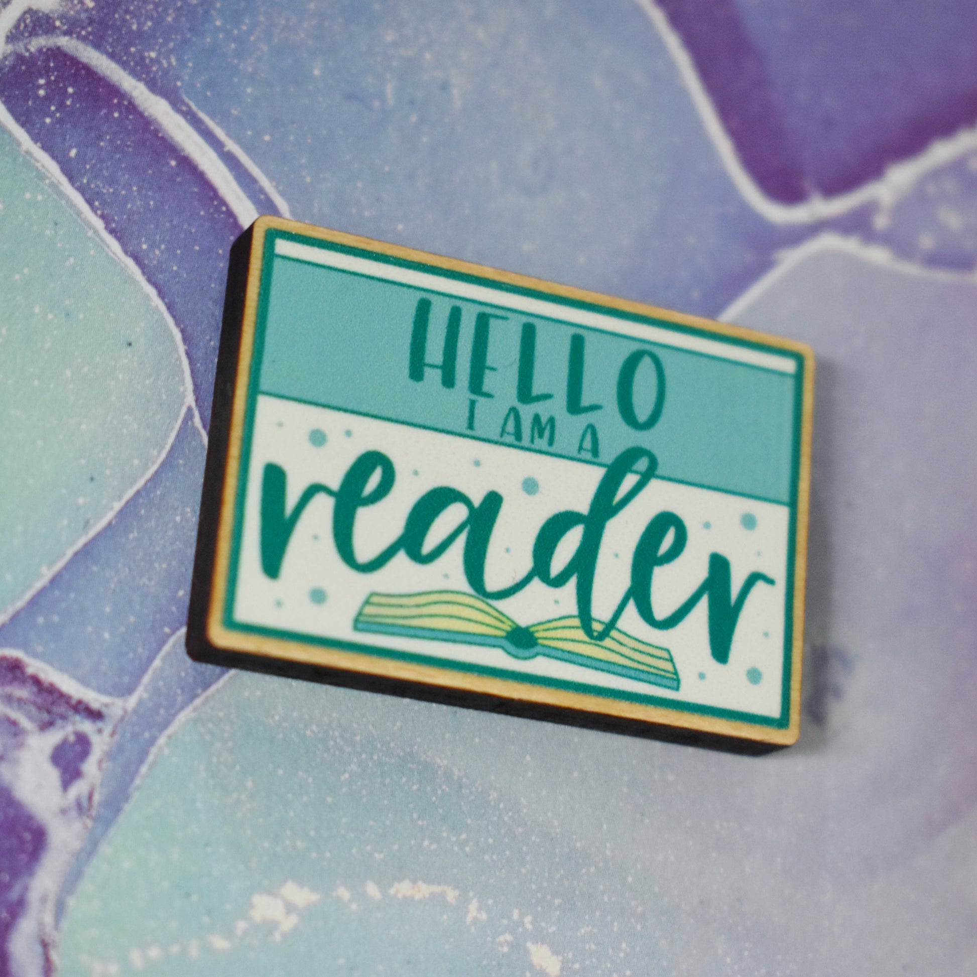 Close up of wooden pin in centre of photo at an angle, which shows the cut edge of the pin which is darker than the wood visable on the front. Design features a teal thin border around the edge, with a lighter teal band across the design, with some white above and below the band. Hello I am a is written in a sans serif font across the tela band. Reader is written in the larger botton white space with an open book beneath. Background is soft watercolour teal and purple style.