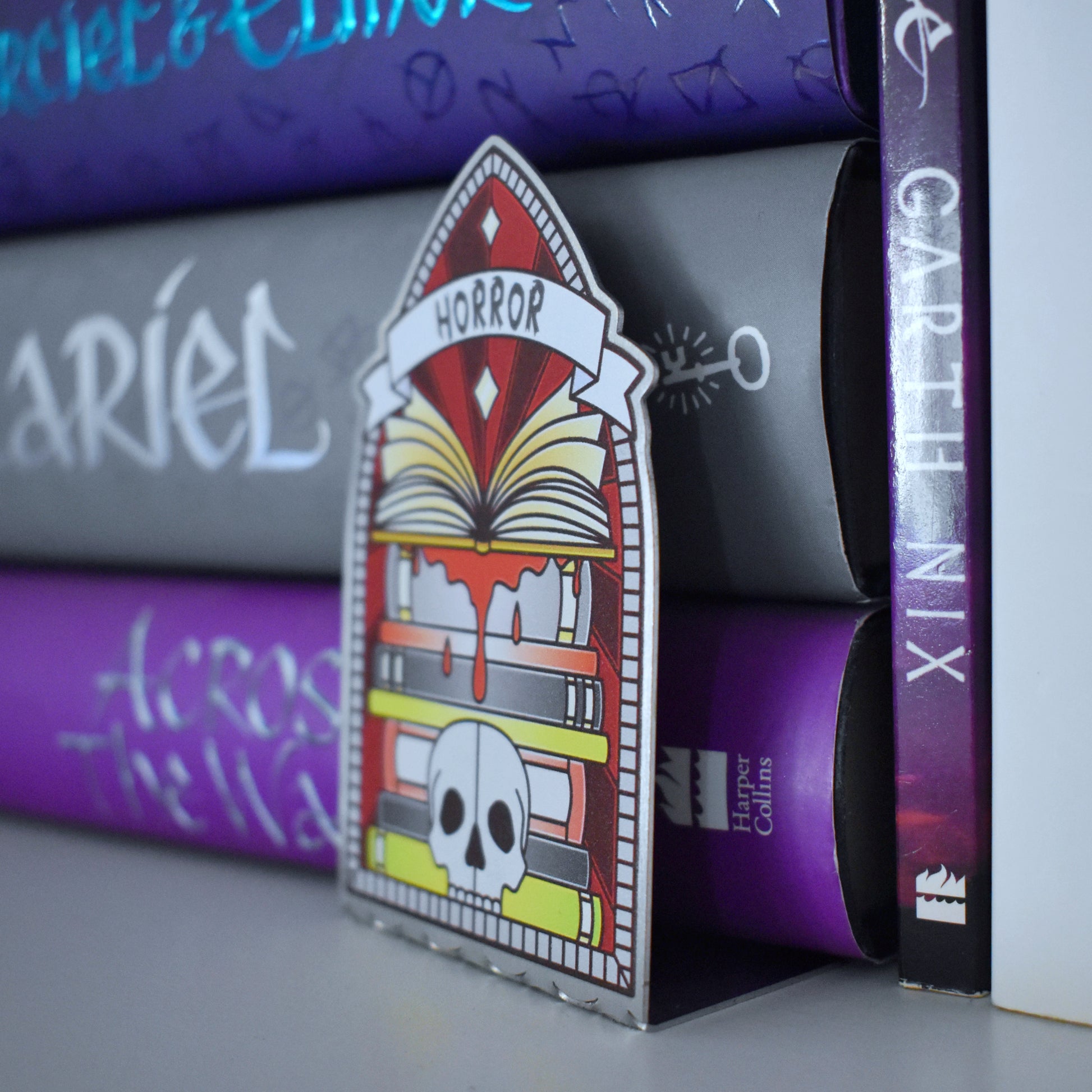 Decorative stainless steel standee with 'Horror' design on in the style of a stained glass window at an angle partically pushed under the book stack, to see it is an L shape that fits under books.