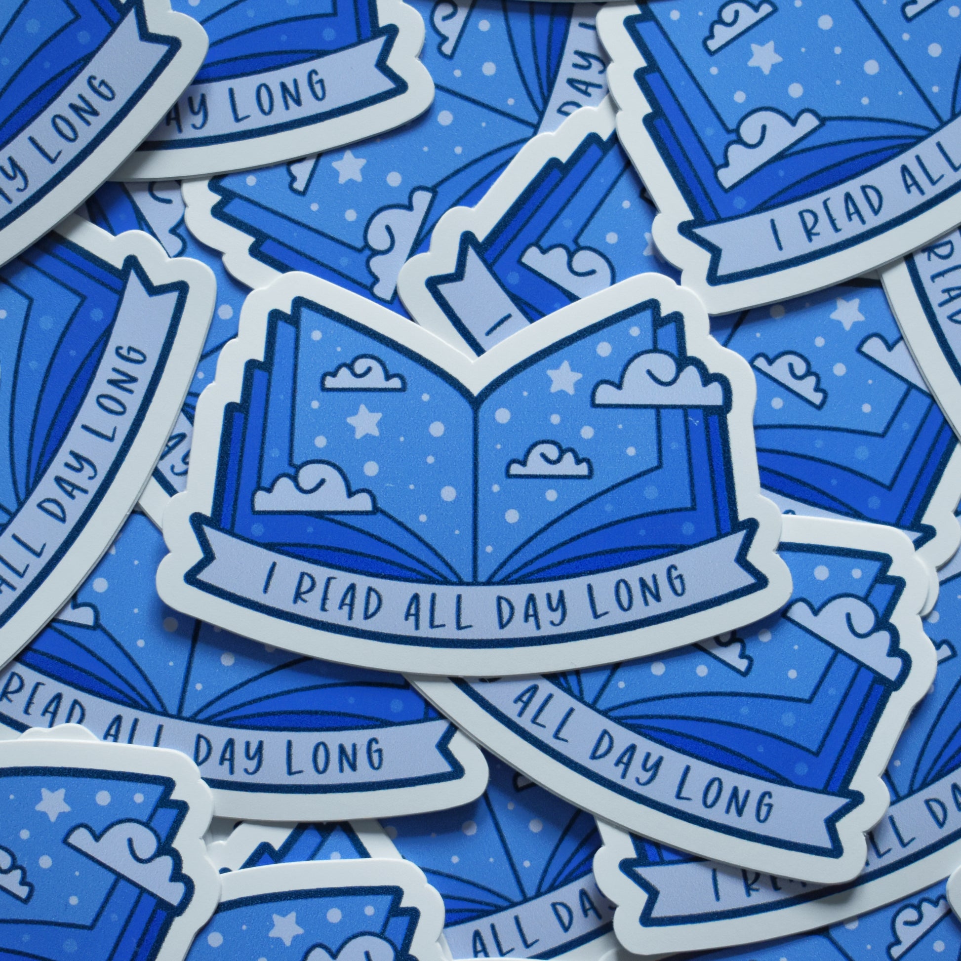 Multiple of same sticker scattered. Sticker design is an open book with a mid to dark blue gradient, with off white/pale blue clouds and pale blue dots/stars spread across the design. An off white/pale blue banner at the bottom reads I read all day long. Sticker has a white border.