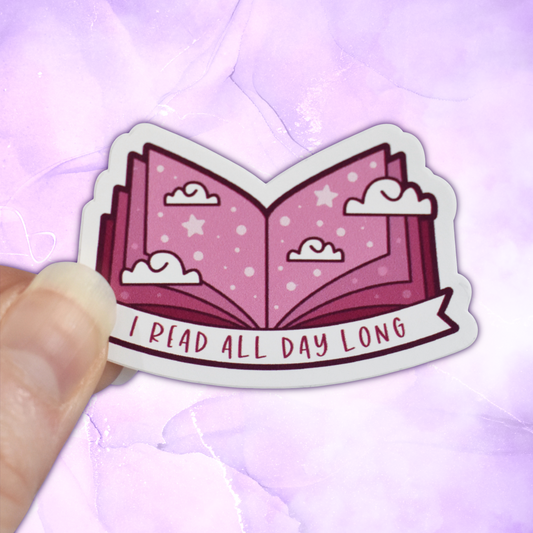 Bookish mini sticker with 'I read all day long' text on a banner, on top of open dusty pink pages book with scattered clouds & stars/dots, held by a white hand against a pastel purple background.