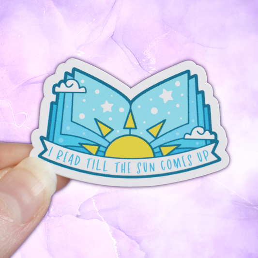 Bookish mini sticker with 'I read till the sun comes up' text on a banner, on top of open light blue pages book with a sun & scattered clouds & stars/dots, held by a white hand against a pastel purple background