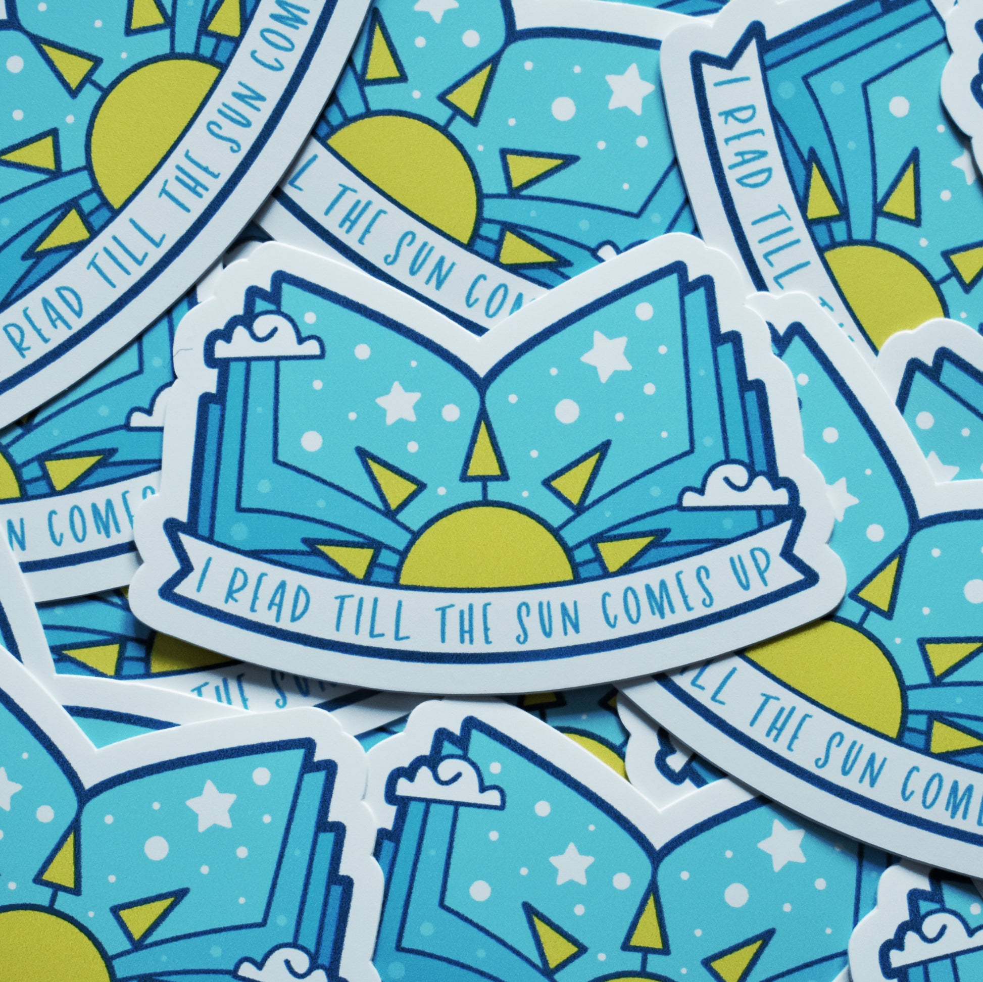 Multiple of same sticker scattered. Sticker design is an open book with a pale blue gradient, with a yellow sun in the middle and white dots/stars/clouds spread across the design. A white banner at the bottom reads I read till the sun comes up. Sticker has a white border.
