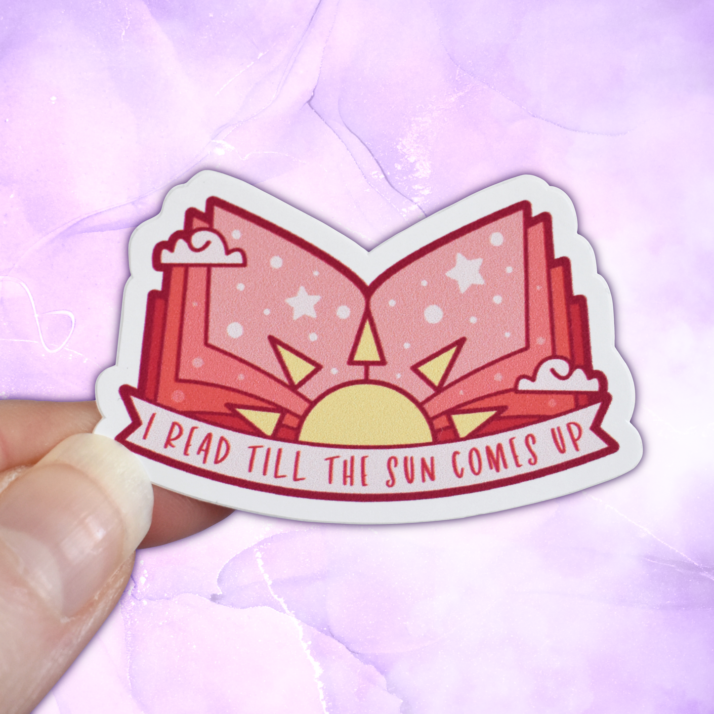 Bookish mini sticker with 'I read till the sun comes up' text on a banner, on top of open peach pages book with a sun & scattered clouds & stars/dots, held by a white hand against a pastel purple background