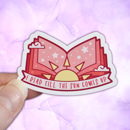 Bookish mini sticker with 'I read till the sun comes up' text on a banner, on top of open peach pages book with a sun & scattered clouds & stars/dots, held by a white hand against a pastel purple background
