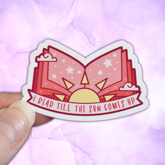 Bookish mini sticker with 'I read till the sun comes up' text on a banner, on top of open peach pages book with a sun & scattered clouds & stars/dots, held by a white hand against a pastel purple background