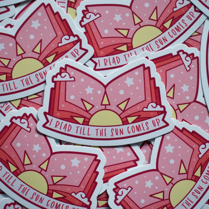 Multiple of same sticker scattered. Sticker design is an open book with a pink/peach gradient, with a yellow sun in the middle and white dots/stars/clouds spread across the design. Off white banner at the bottom reads I read till the sun comes up. Sticker has a white border.
