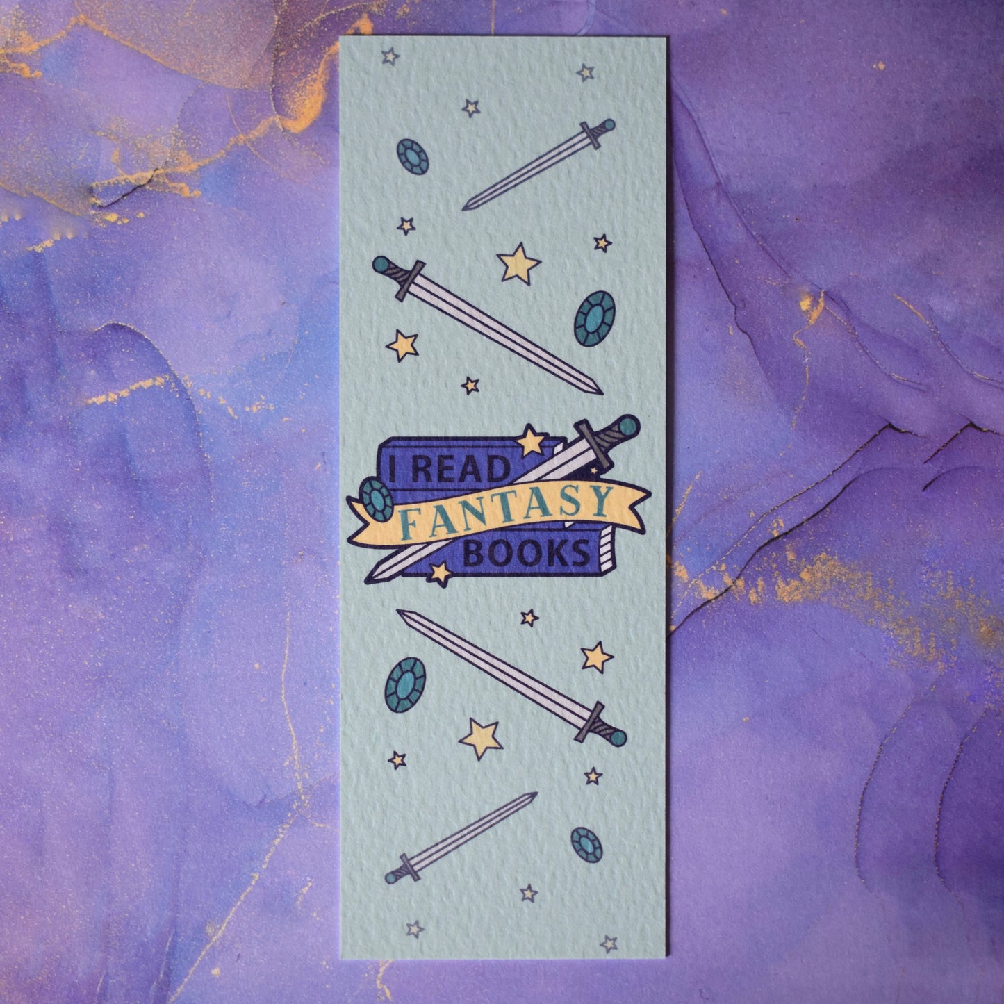 Textured bookmark with 'I Read Fantasy Books' text on a small stack of books and banner, with a sword illustration, in the centre of the design. Smaller swords, gems and stars fill the rest of the bookmark space. Background under bookmark is a purple watercolour.
