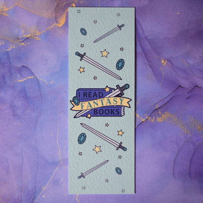 Textured bookmark with 'I Read Fantasy Books' text on a small stack of books and banner, with a sword illustration, in the centre of the design. Smaller swords, gems and stars fill the rest of the bookmark space. Background under bookmark is a purple watercolour.