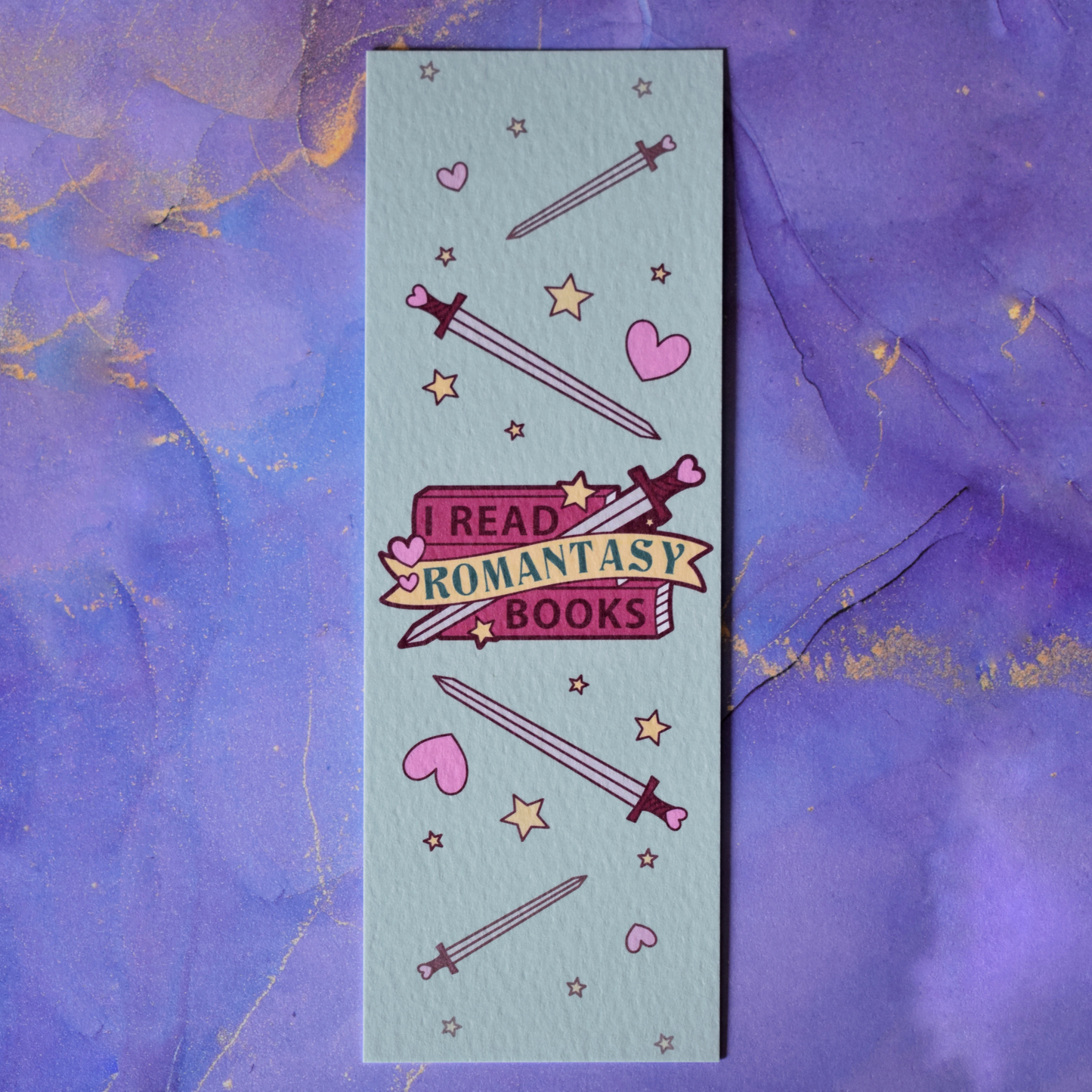 Textured bookmark with 'I Read Romantasy Books' text on a small stack of books and banner, with a sword illustration, in the centre of the design. Smaller swords, hearts and stars fill the rest of the bookmark space. Background under bookmark is a purple watercolour.