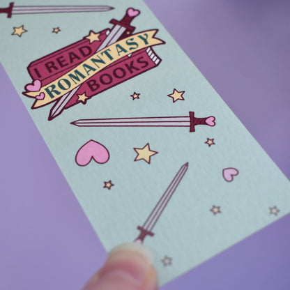 Close up of textured bookmark with 'I Read Romantasy Books' text on a small stack of books and banner, with a sword illustration, in the centre of the design. Smaller swords, hearts and stars fill the rest of the bookmark space. Held by a white hand against lilac background.
