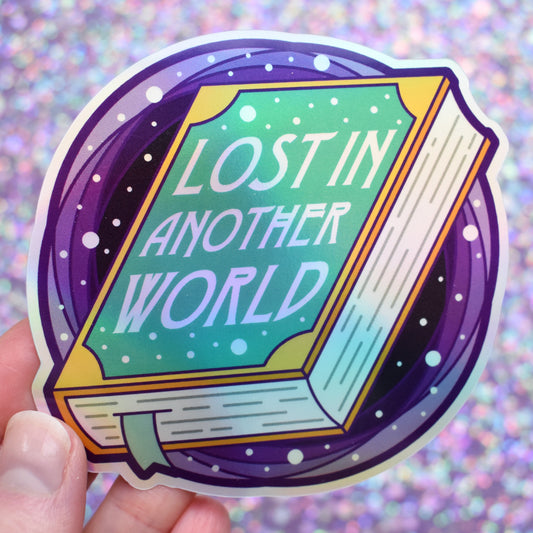 Bookish holographic sticker 'Lost in Another World' text with a closed book in the centre and purple/black hole behind, held by a white hand against a glittery purple background