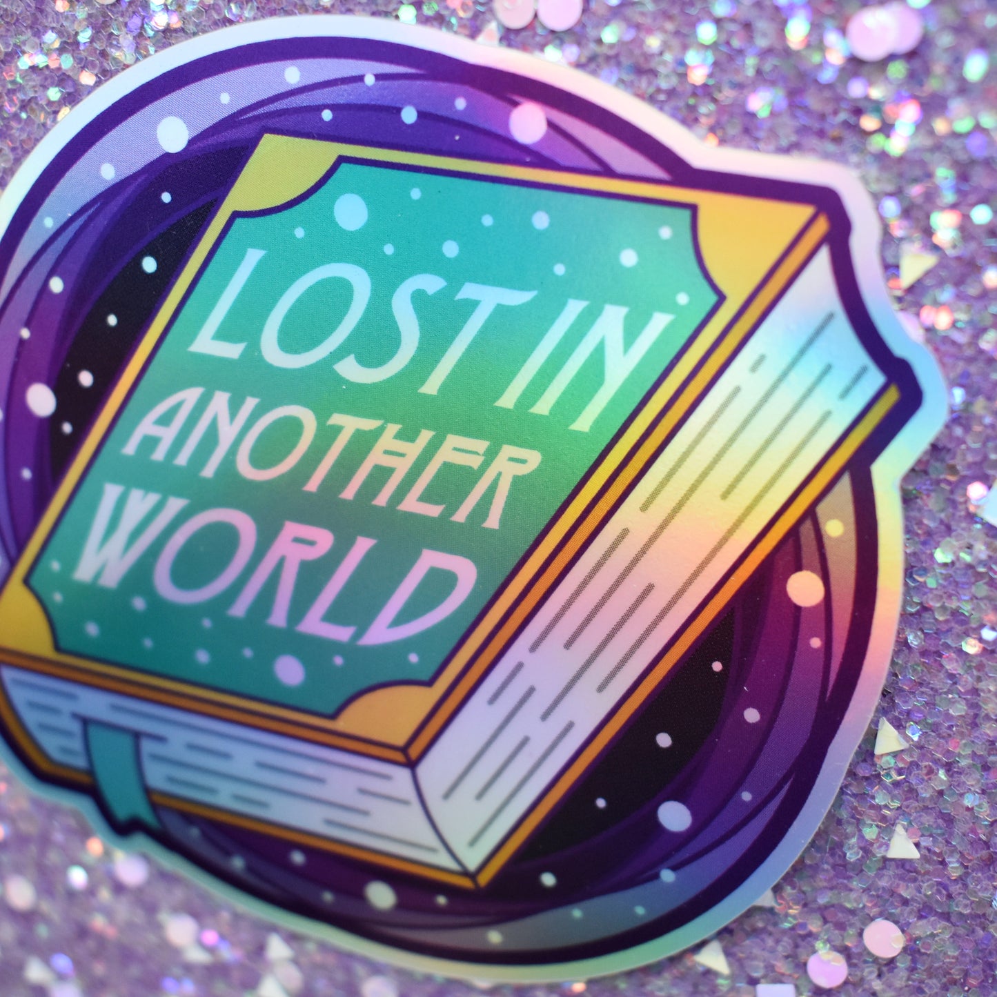 Close up angle of bookish holographic sticker 'Lost in Another World' text with a closed book in the centre and purple/black hole behind, against a glittery purple background