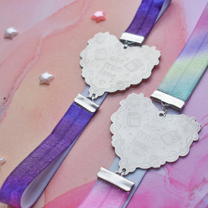 2 book bands at diagonal in the centre of image. Bottom bookband is pastel rainbow elastic with stainless steel engraved heart and book charm that reads one true love. Top bookband is purple galaxy elastic with stainless steel engraved heart and book charm that reads one true love. On a pale pink/peach/lilac watercolour background with different coloured bead stars scattered around.