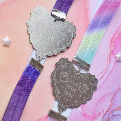 2 book bands at diagonal in the centre of image. Bottom bookband is pastel rainbow elastic with stainless steel engraved heart and book charm that reads one true love. Top bookband is purple galaxy elastic with stainless steel engraved heart and book charm that reads one true love. On a pale pink/peach/lilac watercolour background with different coloured bead stars scattered around.
