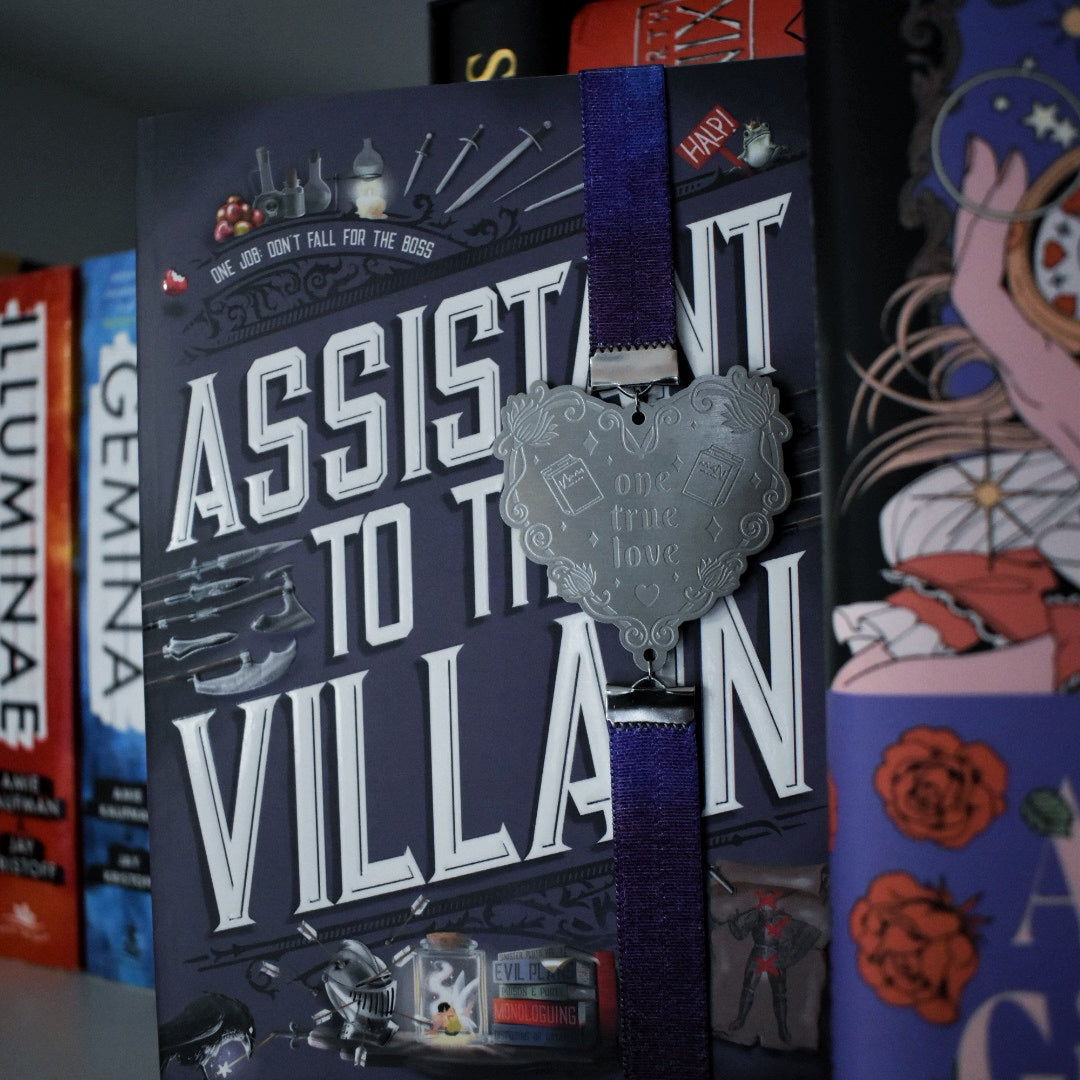 Book titled Assistant to the Villain is shown with a book band wrapped arpound top to bottom on right hand side. The bookband is purple galaxy elastic with stainless steel engraved heart and book charm that reads one true love. Bookshelf and other books can be seen in background and forground of image.