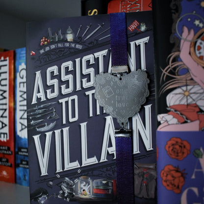 Book titled Assistant to the Villain is shown with a book band wrapped arpound top to bottom on right hand side. The bookband is purple galaxy elastic with stainless steel engraved heart and book charm that reads one true love. Bookshelf and other books can be seen in background and forground of image.