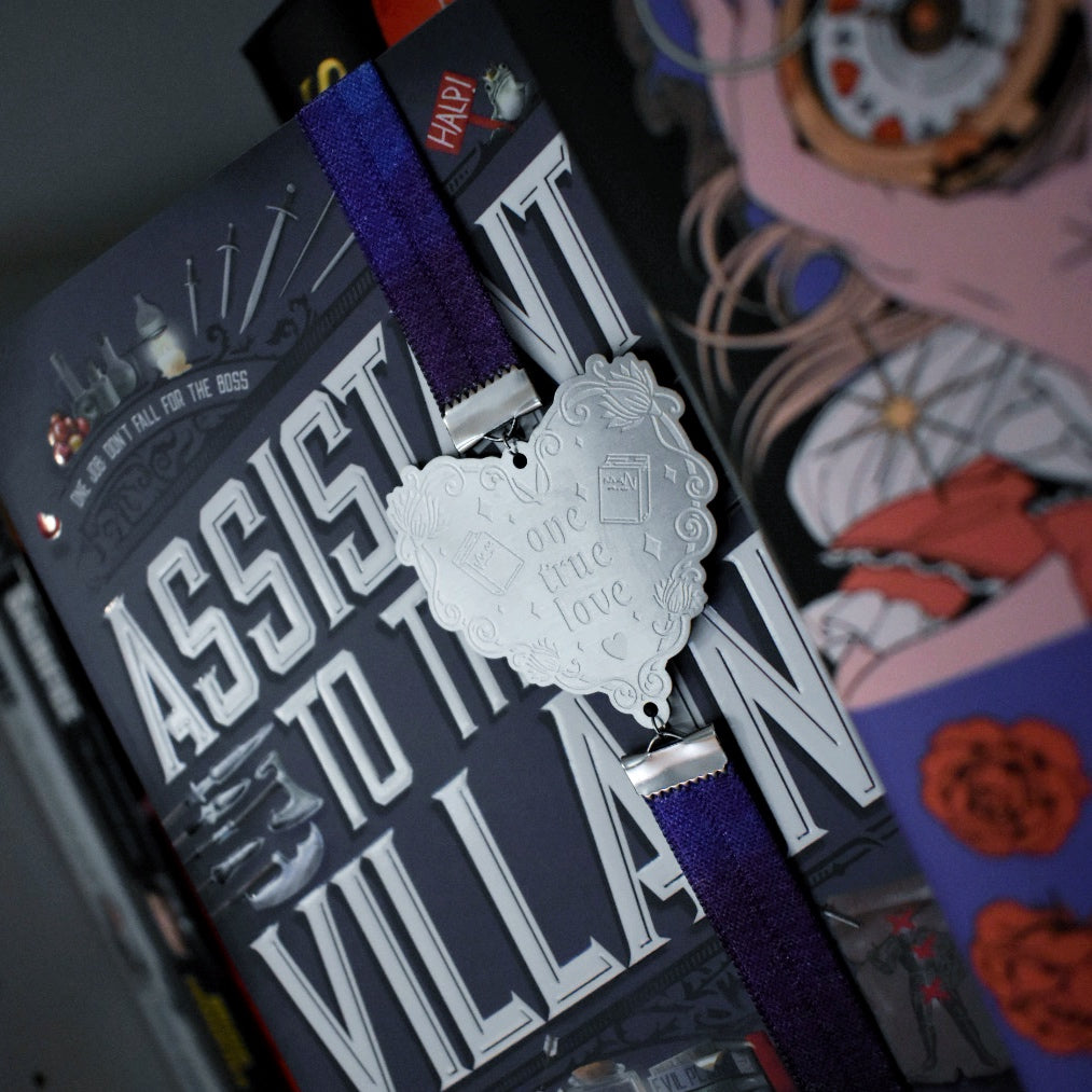 Book titled Assistant to the Villain is shown with a book band wrapped arpound top to bottom on right hand side. The bookband is purple galaxy elastic with stainless steel engraved heart and book charm that reads one true love. Bookshelf and other books can be seen in background and forground of image.