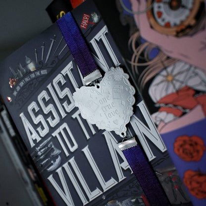 Book titled Assistant to the Villain is shown with a book band wrapped arpound top to bottom on right hand side. The bookband is purple galaxy elastic with stainless steel engraved heart and book charm that reads one true love. Bookshelf and other books can be seen in background and forground of image.