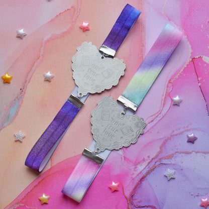 2 book bands at diagonal in the centre of image. Bottom bookband is pastel rainbow elastic with stainless steel engraved heart and book charm that reads one true love. Top bookband is purple galaxy elastic with stainless steel engraved heart and book charm that reads one true love. On a pale pink/peach/lilac watercolour background with different coloured bead stars scattered around.