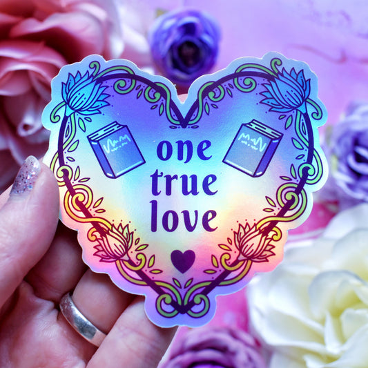 Purple heart-shaped bookish holographic sticker design features text 'one true love' with a floral and swirly outline with book covers either side of the text. Held by a white hand above a watercolour style background in blue, pink and purples with flowers around the edge of the photo.
